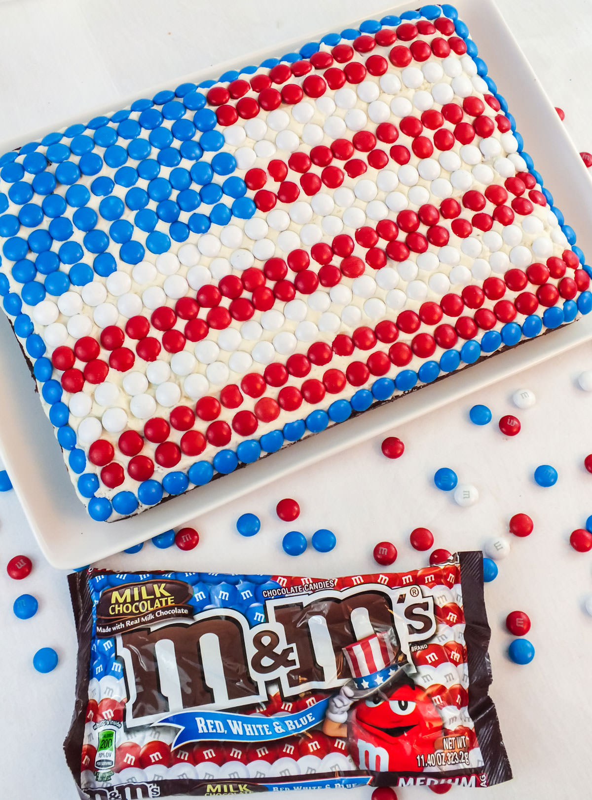 Closeup of an American Flag Cake sitting on a white cake platter on a white table next to a bag of Red White and Blue M&M's.