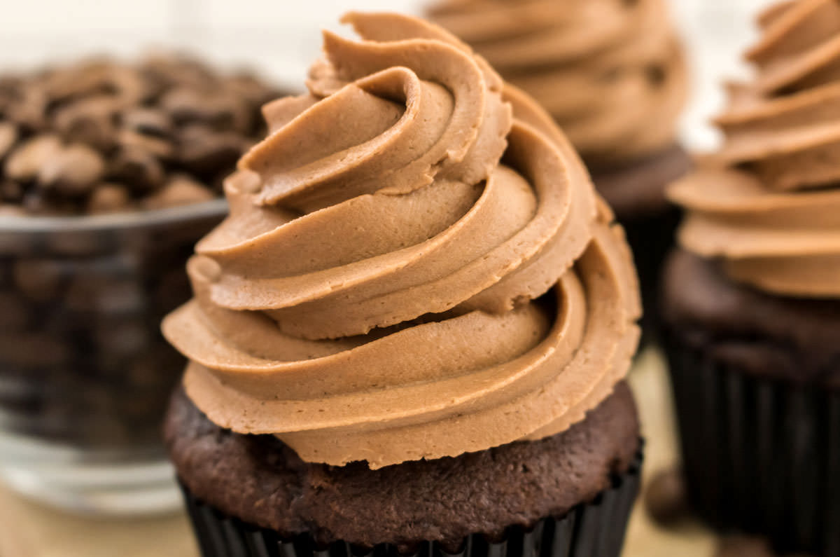Closeup on three cupcakes topped with The Best Mocha Buttercream Frosting sitting in front of a glass bowl filled with coffee beans.