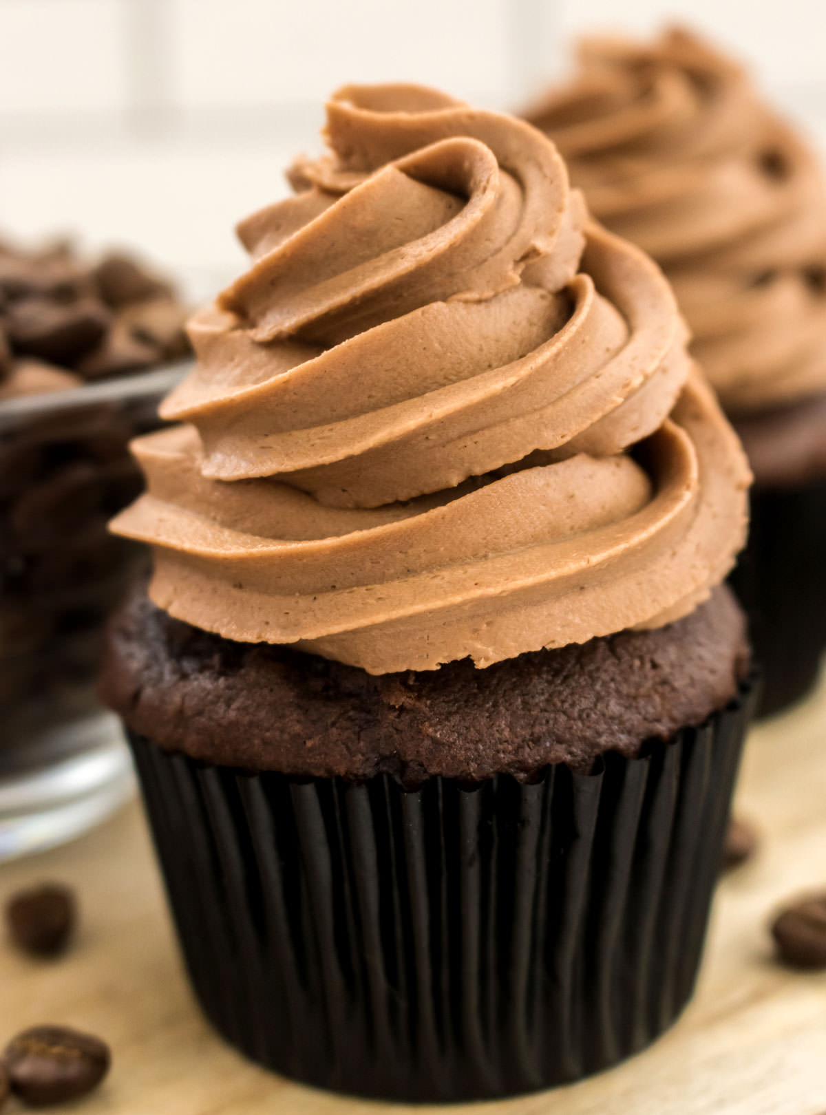 Closeup on a chocolate cupcake topped with The Best Mocha Buttercream Frosting sitting on a cutting board next to a glass bowl filled with coffee beans.