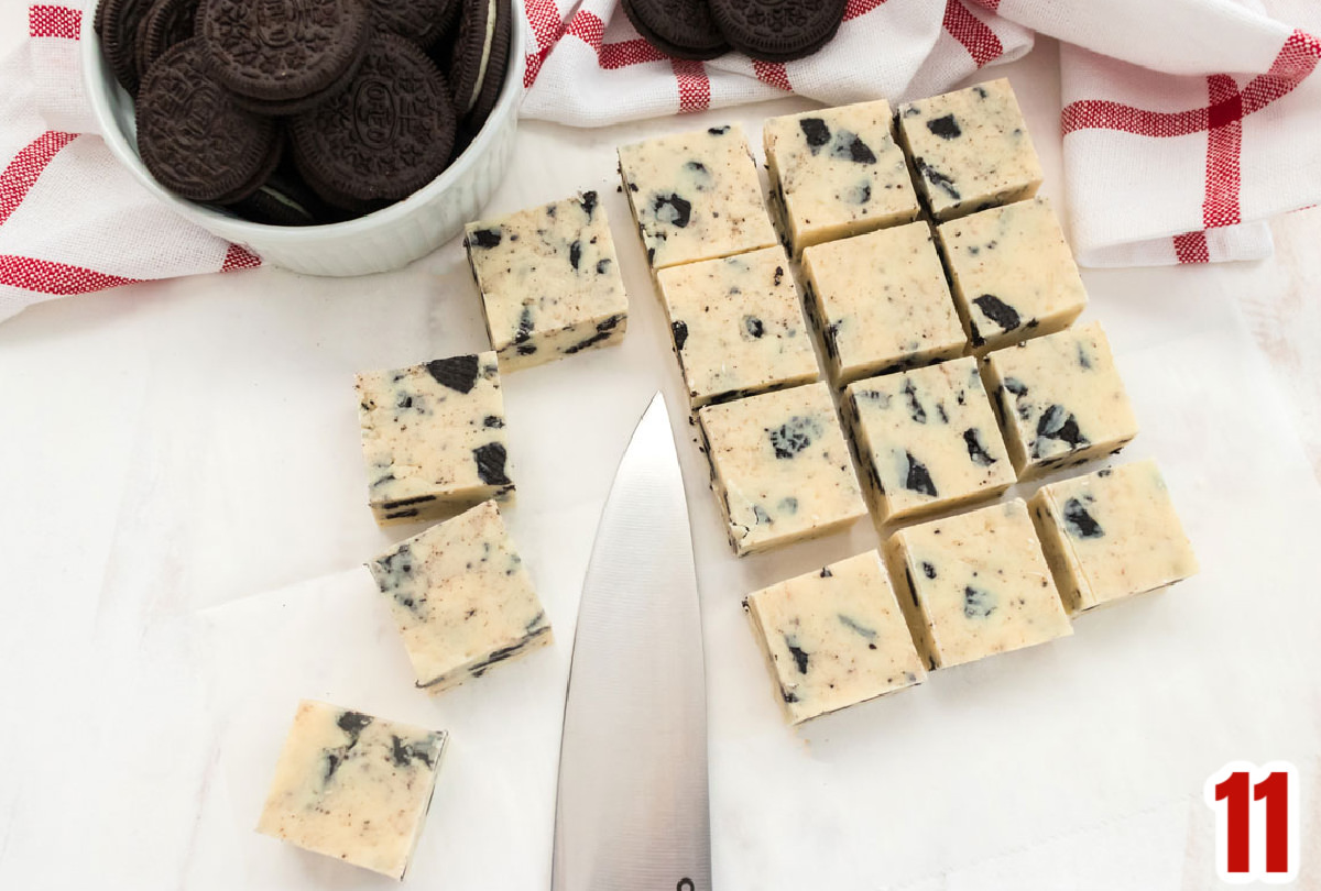 Sixteen pieces of Oreo Fudge laying on white surface in rows with a knife and a ramekin filled with Oreo Cookies.