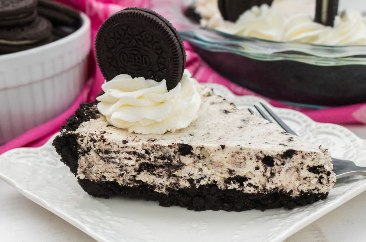 Closeup on a side-view of the Oreo Pie sitting on a white plate on a white table with pink linens in front of a ramekin filled with Oreo Cookies.