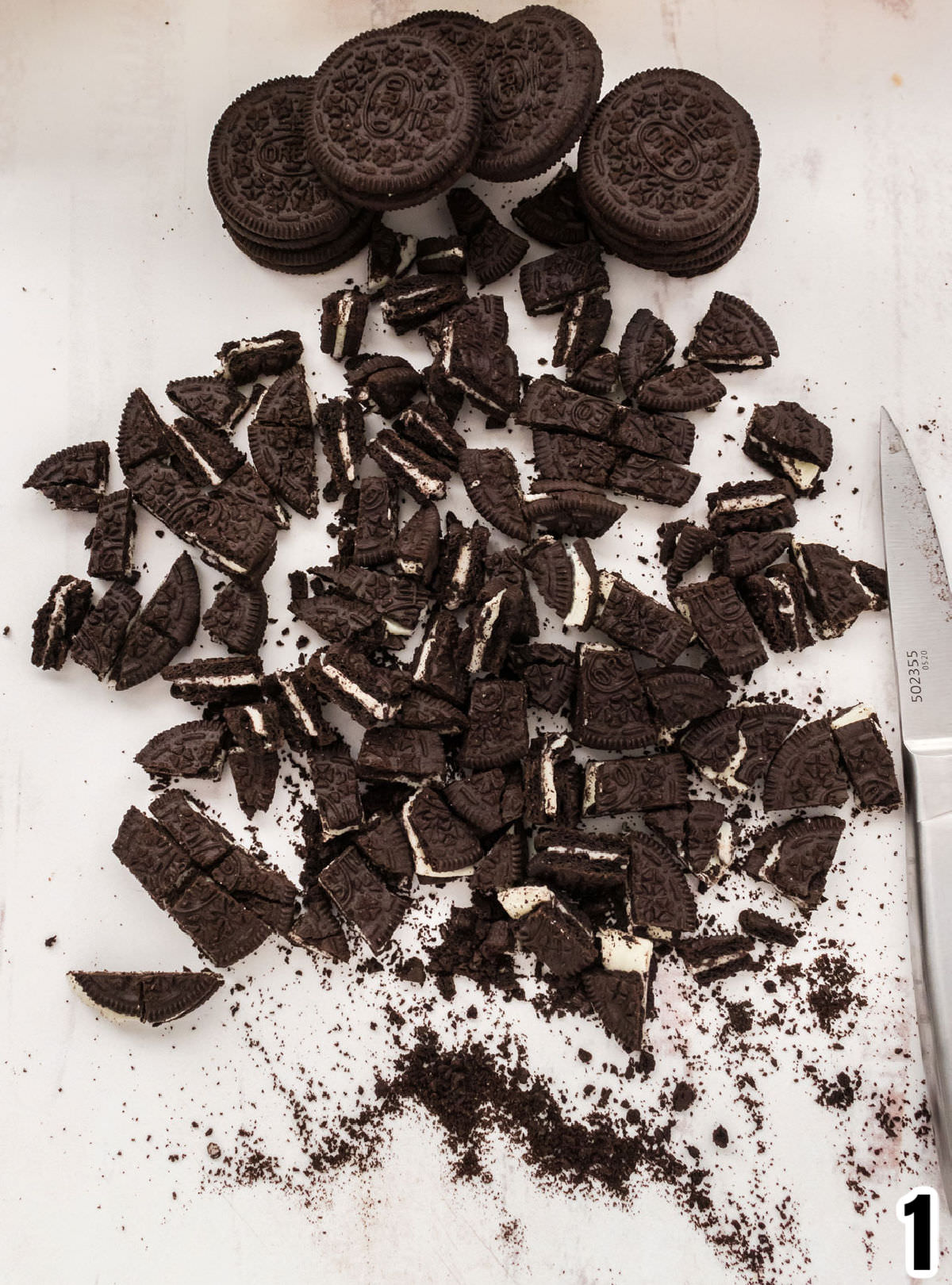 Oreo Cookies, some in pieces, some whole, on a white surface with a silver kitchen knife.