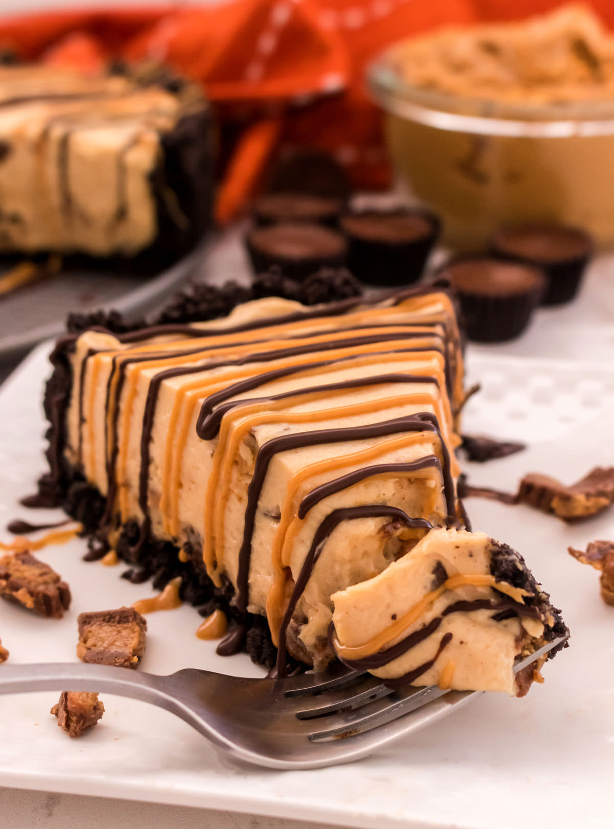 Closeup on a single slice of No Bake Peanut Butter Cheesecake sitting on a white plate in front of a bowl of peanut butter, the rest of the cheesecake and Mini Reeses Peanut Butter Cups.