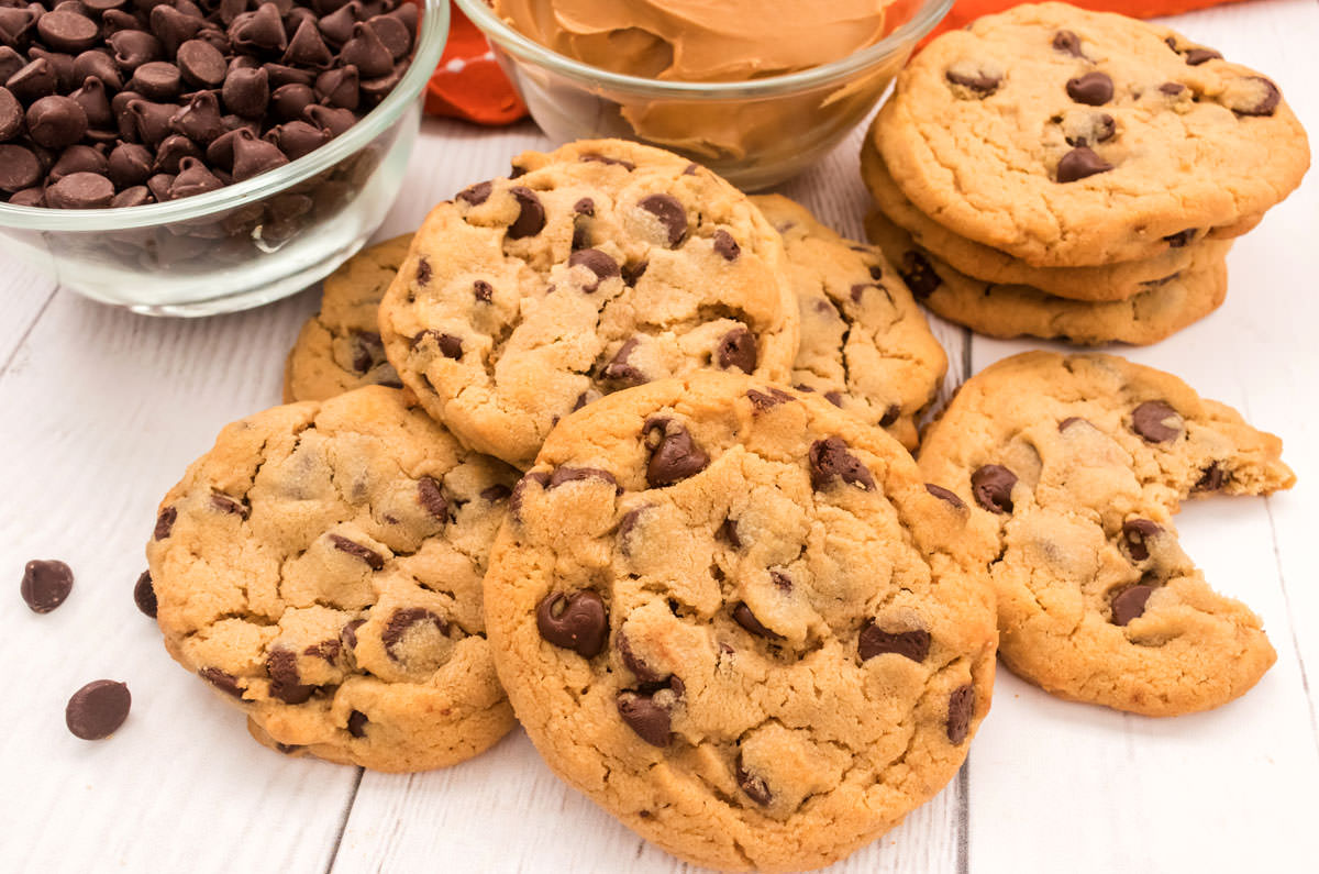 Closeup on a batch of Peanut Butter Chocolate Chip Cookies laying on a white table in front of ramekins filled with chocolate chips and peanut butter.