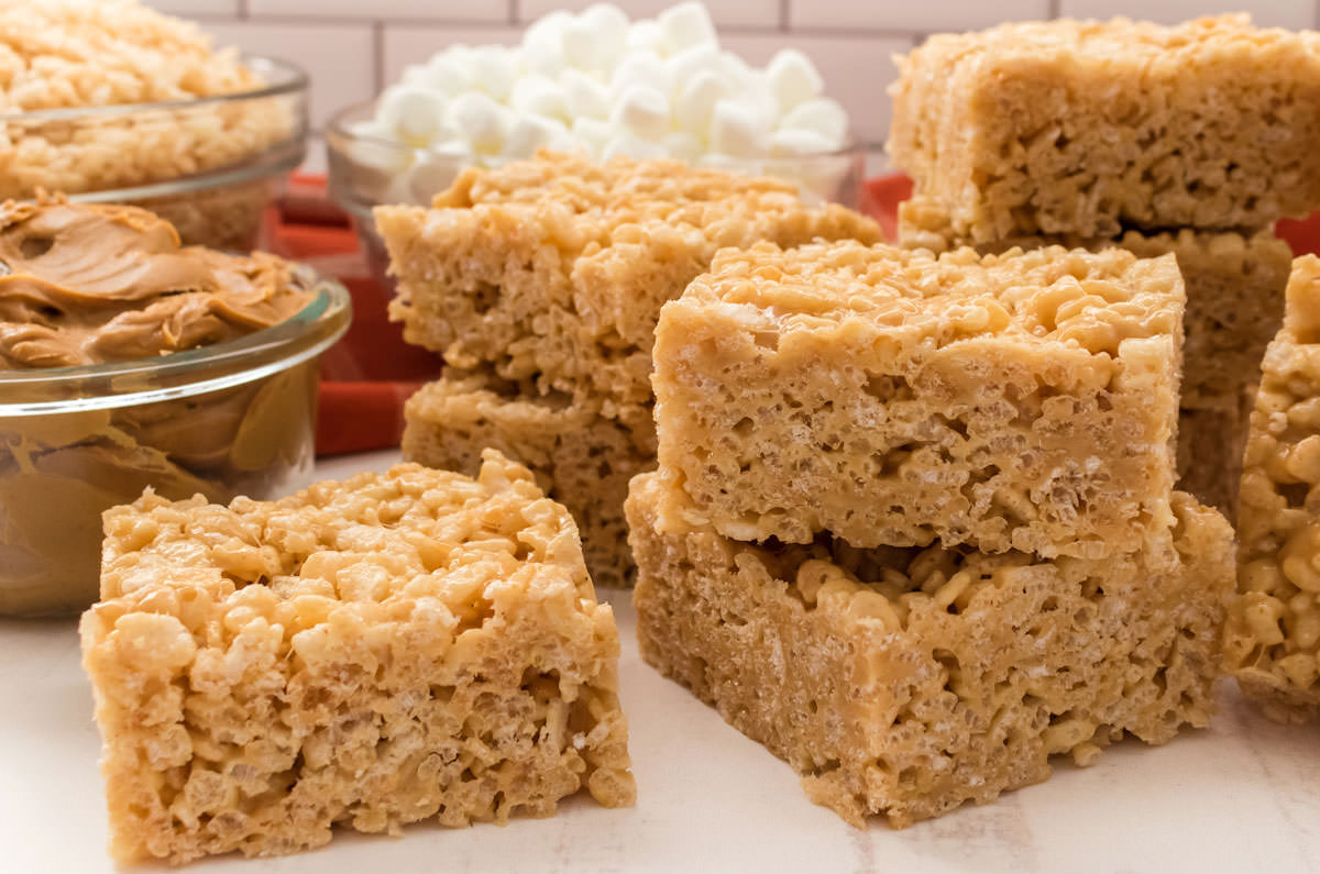 Closeup on a batch of Peanut Butter Rice Krispie Treats sitting on a white surface surrounded by cereal, marshmallows and peanut butter.