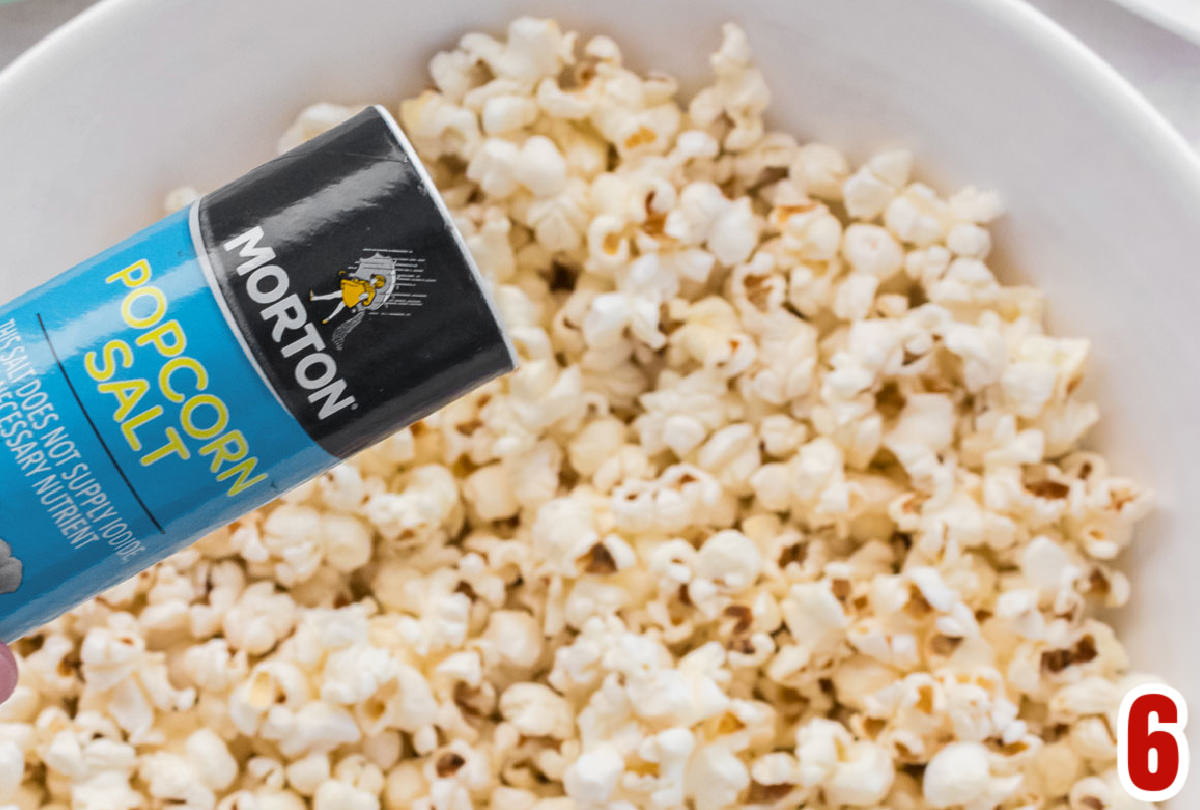 Closeup on a bowl of popcorn with a shaker of Morton Popcorn Salt sprinkling salt on the popcorn.