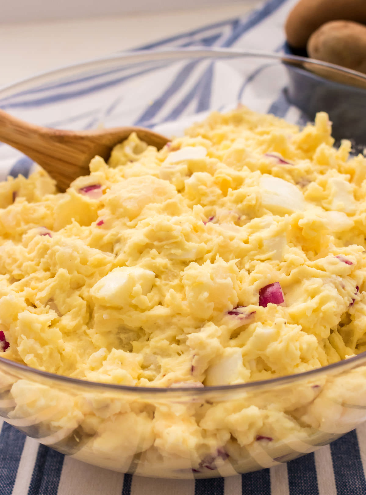 Clear salad bowl filled with Classic Homemade Potato Salad sitting on a blue and white striped kitchen towel.