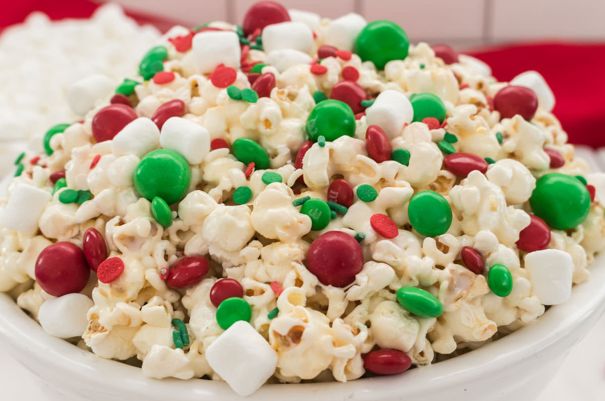 Closeup on a white serving bowl filled with Santa Crunch Popcorn sitting on a white table in front of a bowl of marshmallows and a red towel.