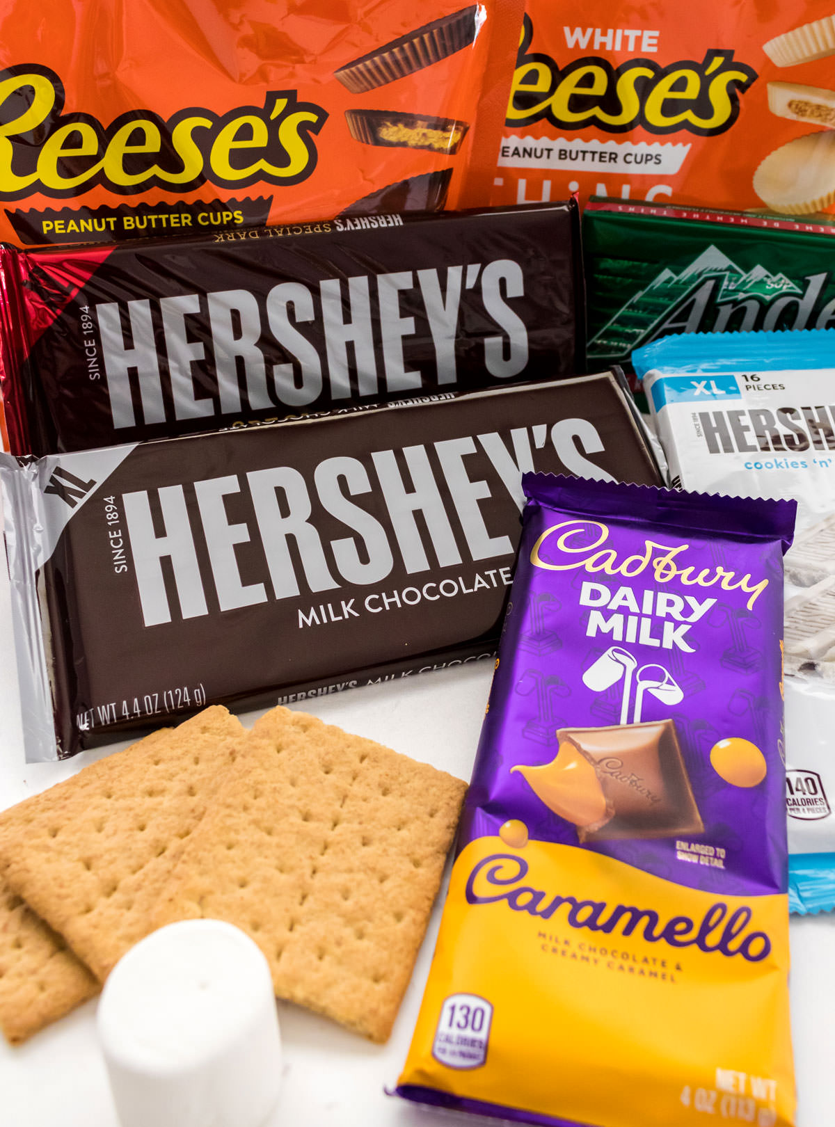 Many types of chocolate candy bars laying on a white table along side graham crackers and a marshmallow.