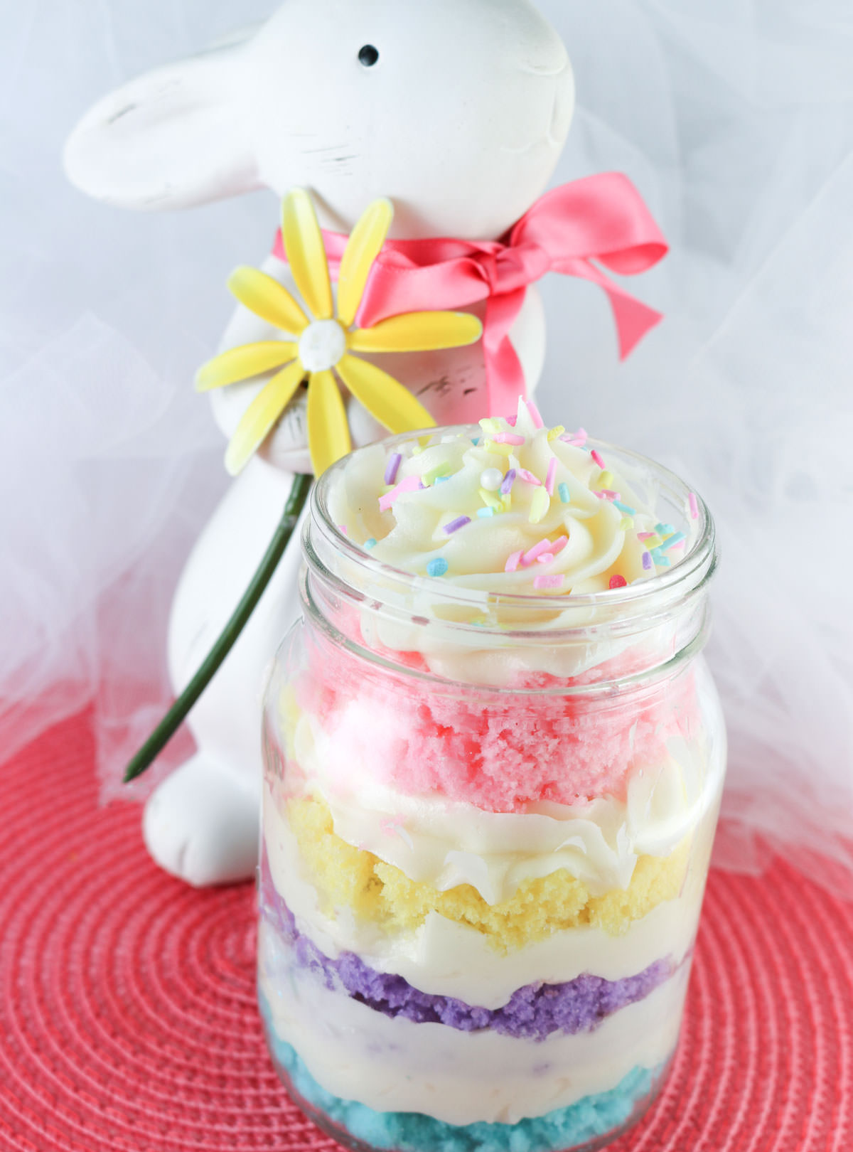 Closeup on a Springtime Cupcake in a  Jar sitting on a pink placemat in front of a white easter bunny decoration holding a yellow flower.