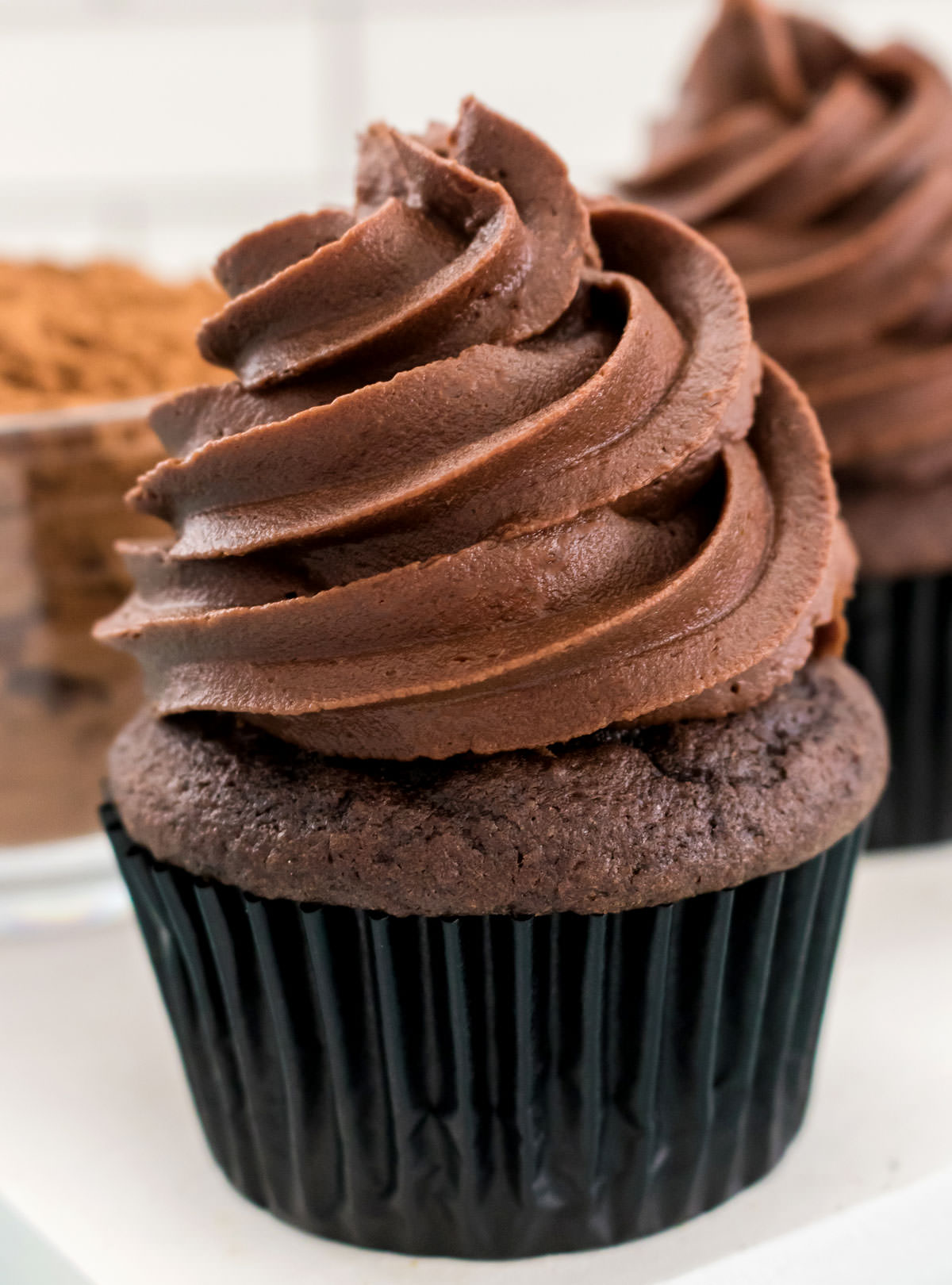 Closeup on a chocolate cupcake topped with a swirl of The Best Chocolate Buttercream Frosting sitting in front of a glass bowl filled with cocoa powder.