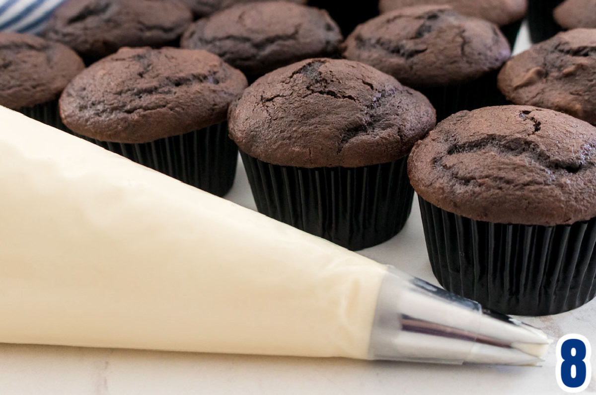 A frosting decorating bag filled with Cream Cheese Frosting sitting on a white table in front of a batch of unfrosted chocolate cupcakes.