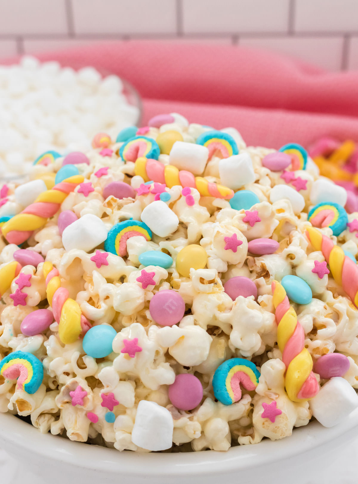 Closeup on a white serving bowl filled with colorful Unicorn Popcorn with a bowl of mini marshmallows in the background and a pink table linen.