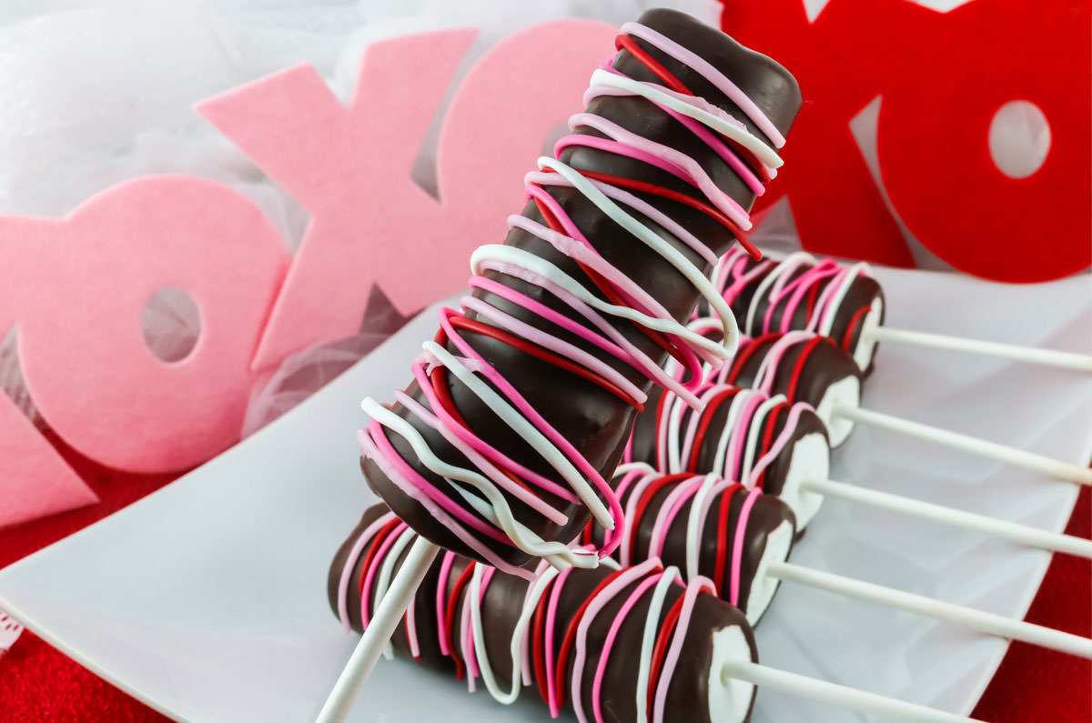 A Valentine's Marshmallow Pop held over a white serving platter holding more chocolate covered marshmallow pops.