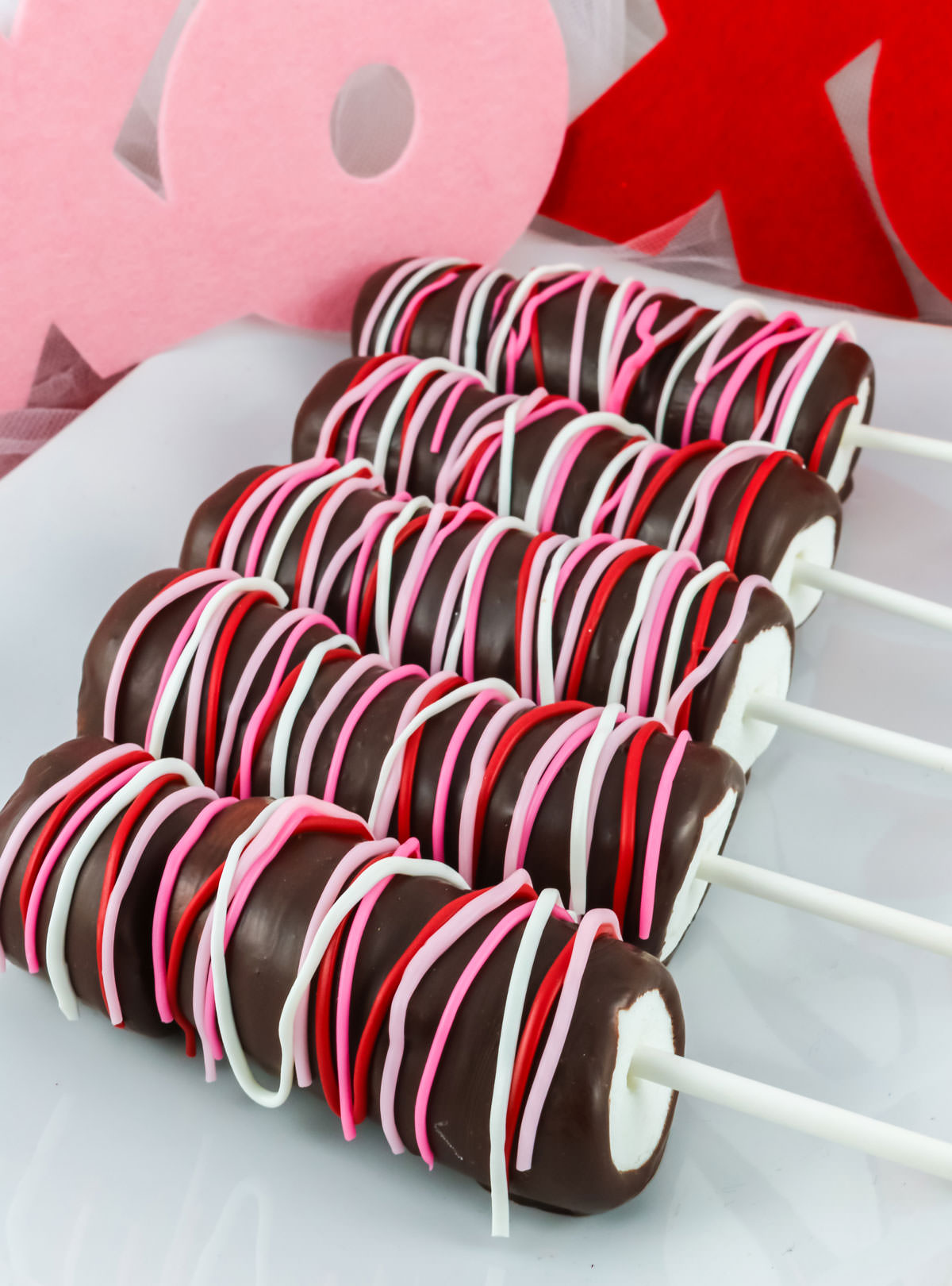 5 Valentine's Marshmallow Pops laying on a white serving platter in from of an XOXO Valentine's Day Decoration.