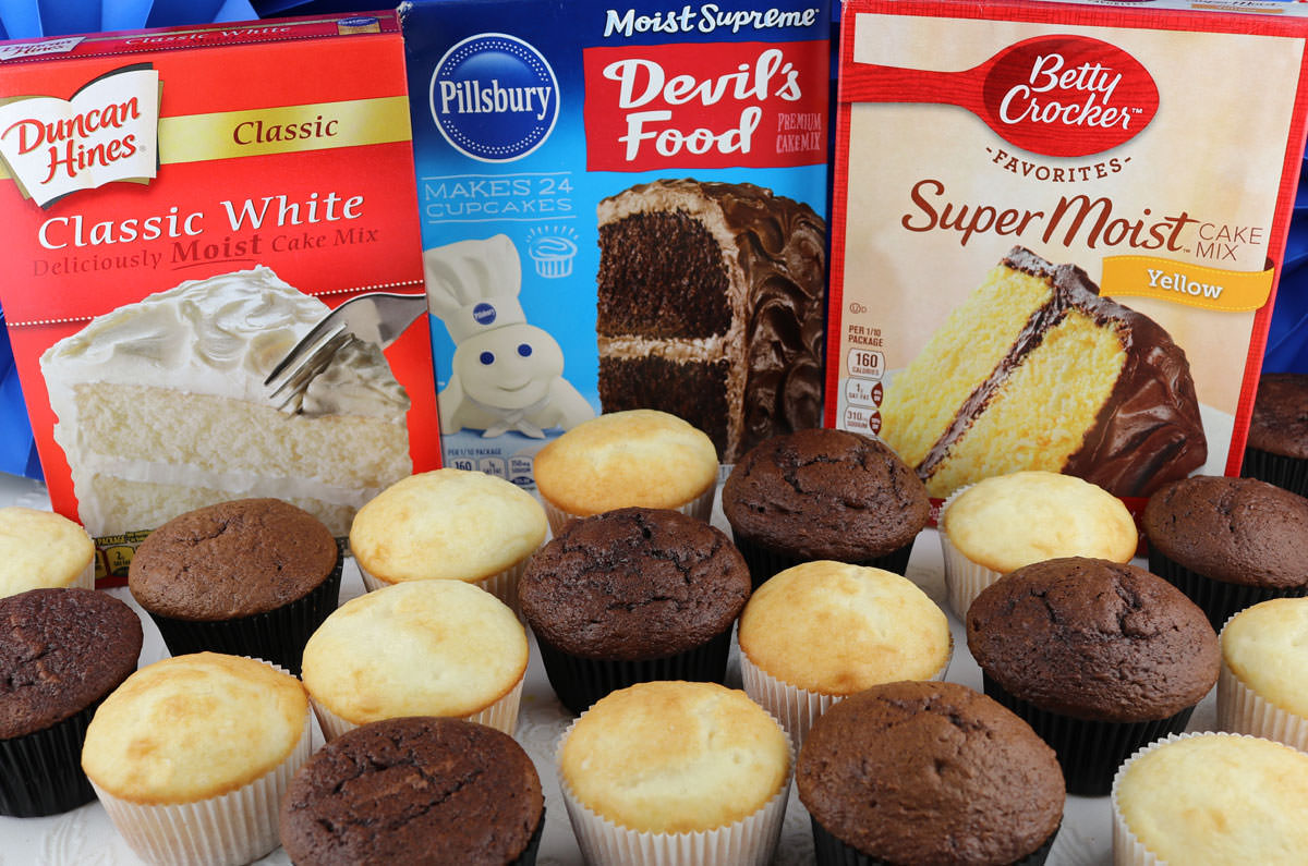 Unfrosted chocolate and vanilla cupcakes sitting on a white table in front of boxes of popular store-bought cake mixes.