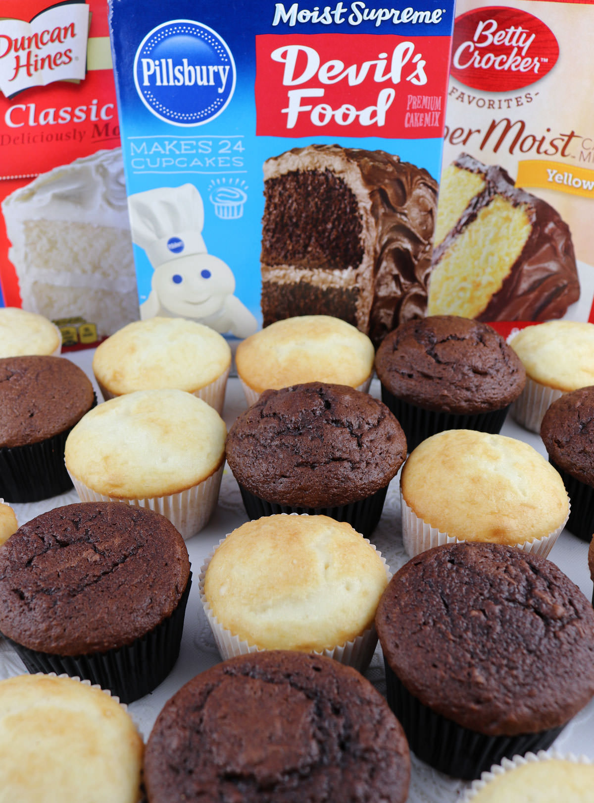 Unfrosted chocolate and vanilla cupcakes sitting on a white table in front of boxes of popular store-bought cake mixes.